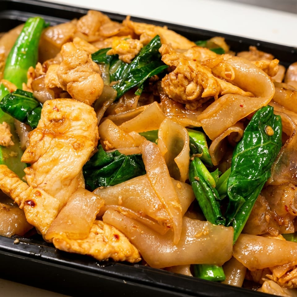 Pad See Ew with wide rice noodles, chicken, and Chinese broccoli in a black takeout container.